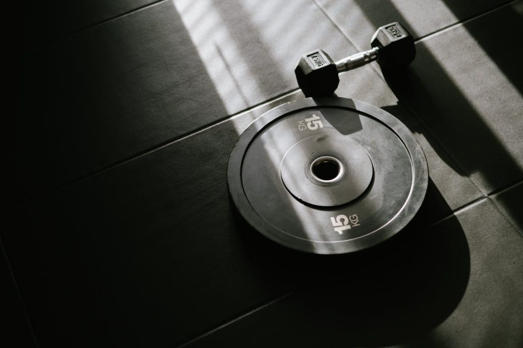pexels-photo-16513600 A 15 kg weight plate and dumbbell on a gym floor, lit by sunlight through a window.